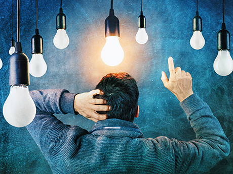 A man is standing with his back to the viewer, scratching his head with his left hand and pointing in the air with his right index finger. Above him hang many light bulbs. One of them is lit up.